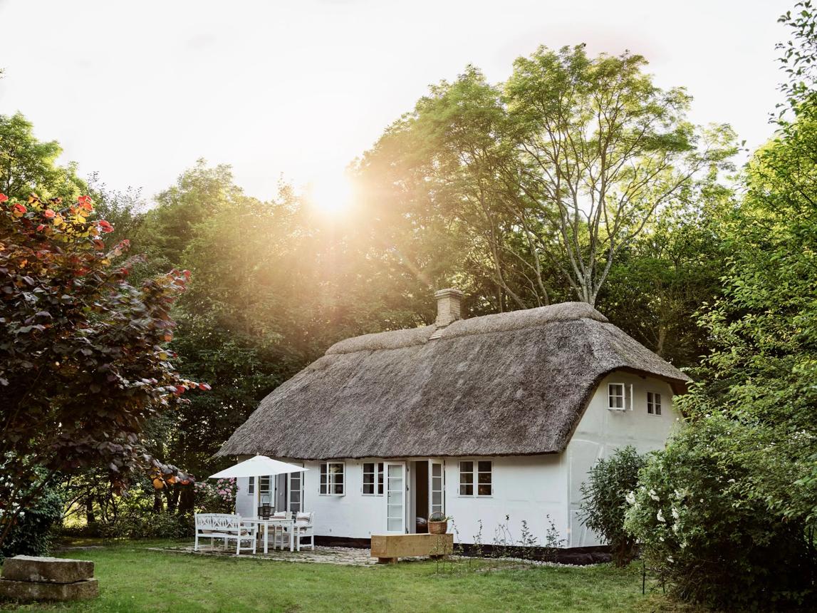 Et hvidt stråtækt hus i en have med aftensol gennem træerne.